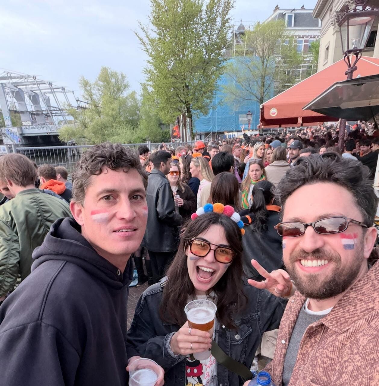 Three friends taking a selfie in the King's Day crowd in Amsterdam, Dutch flags painted on their cheeks