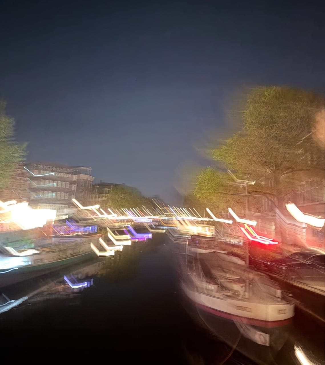 A long-exposure night view of an Amsterdam canal, moored boats and street lights streaking across the water